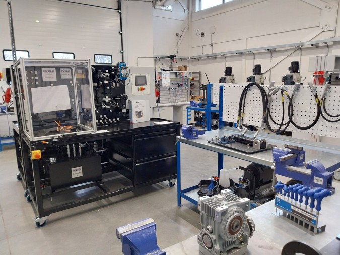 A bright, organized workshop with various industrial machines, workbenches, blue vises, and hydraulic or mechanical equipment arranged neatly throughout the space.