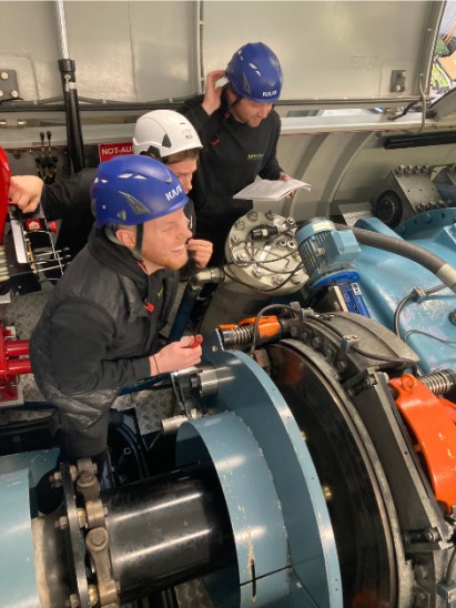 Three men in safety helmets examining a machine inside a wind turbine nacelle.