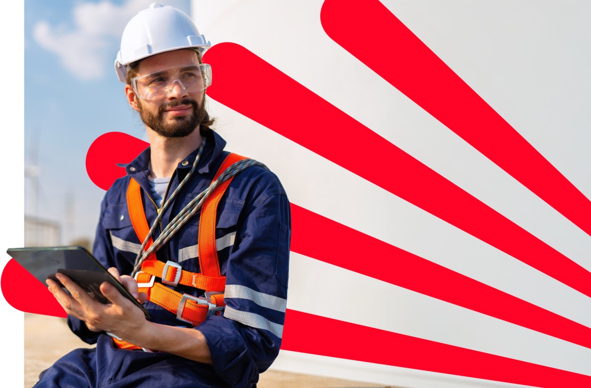 A wind turbine technician in a hard hat and safety vest holds a tablet at a wind farm, overseeing operations.