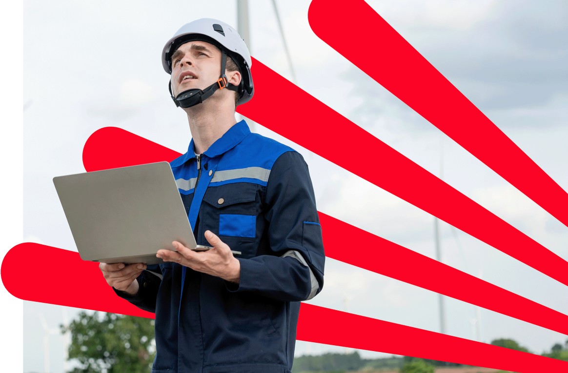 A wind turbine technician in a hard hat and safety glasses holds a laptop on a wind farm, overseeing operations.