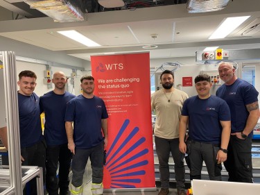 An image of 5 men in blue t-shirts and 1 man in a sand coloured t-shirt standing between a red roller banner that advertises WTS services and how they are changing the status quo.