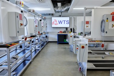 A spacious electrical workshop featuring several control panels, training rigs, and a TV displaying the WTS brand.