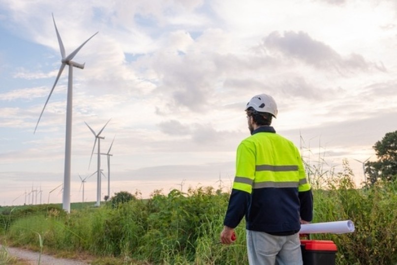 Wind technician in high-vis jacket and helmet standing on a rural track, looking out at a line of wind turbines on the horizon.
