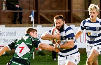 Dom playing rugby holding the ball, with a team mate behind and the opposing player attacking.